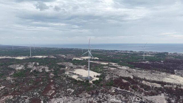 Aerial side perspective of a tall wind turbine with red-tipped blades, rising above the flat coastal terrain with scattered vegetation and the sea beyond.