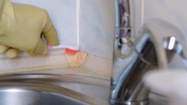 Gloved hand gentle scrubbing discolored grout seam by sink with soft toothbrush, closeup showing light strokes removing brown film from tile joint, domestic renovation and care