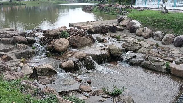 A small rocky stream with cascading water flowing over smooth boulders in a natural outdoor setting. The scene features clear running water, weathered rocks, patches of green vegetation, driftwood, an