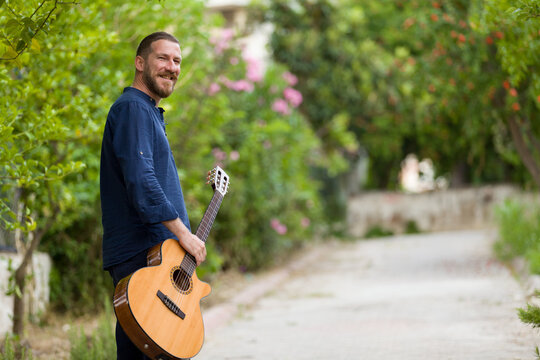 Portrait of a middle-aged bearded man walking through a lush garden with an acoustic classical guitar, looking back with a smile, natural bokeh background.