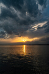 Fototapeta premium Dramatic sunset over Lake Constance seen from the Austrian shore in Bregenz, Vorarlberg, with the golden sun breaking through dark storm clouds and reflecting on the calm water surface