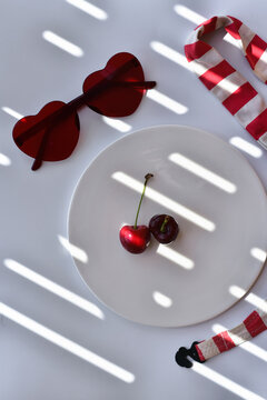 Red heart shaped sunglasses, cherries on a plate in the shade on white background