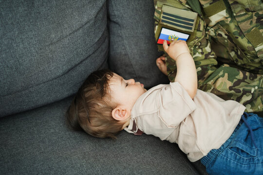 Male russian soldier in military uniform hugs his little son at home while baby boy touching russian flag - Emotional military happy homecoming concept - Main focus on kid head