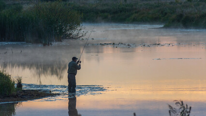 fisherman stands in the calm, misty river during a breathtaking golden sunrise, holding his rod and patiently waiting for a catch. © Александр Арендарь