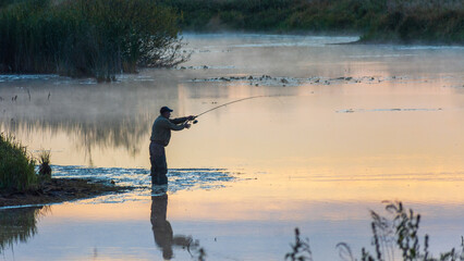 fisherman stands in the calm, misty river water during a peaceful sunrise, casting his line into the golden reflection © Александр Арендарь