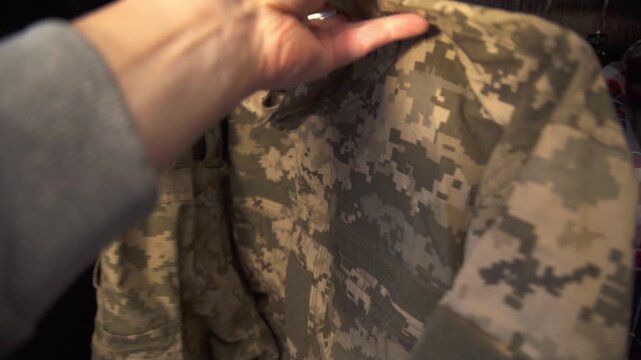 A man touches casual shirts before taking a military camouflage jacket out of a closet.