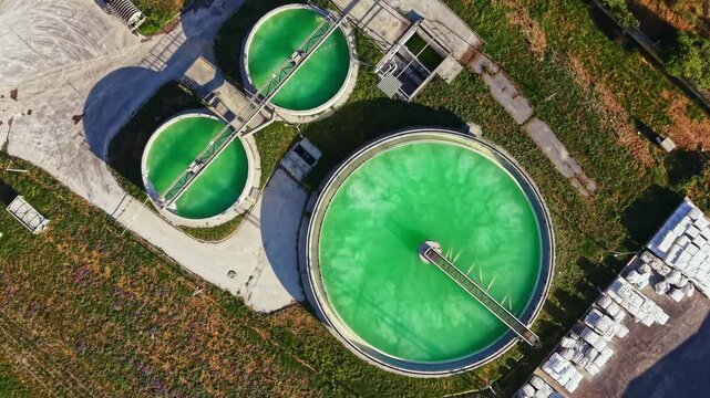 Three circular tanks are filled with green liquid in a wastewater treatment facility. The tanks are surrounded by grass and trees under clear blue sky, taken from above.