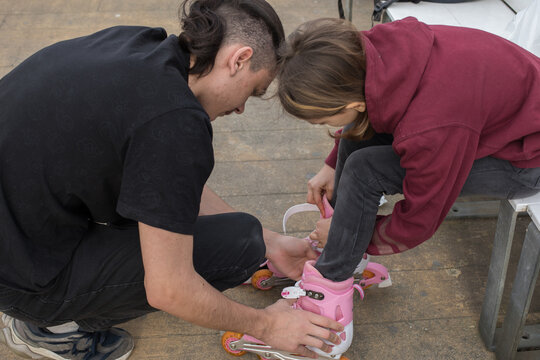 A child sits on a bench in a park while an adult roller skates on their feet.
