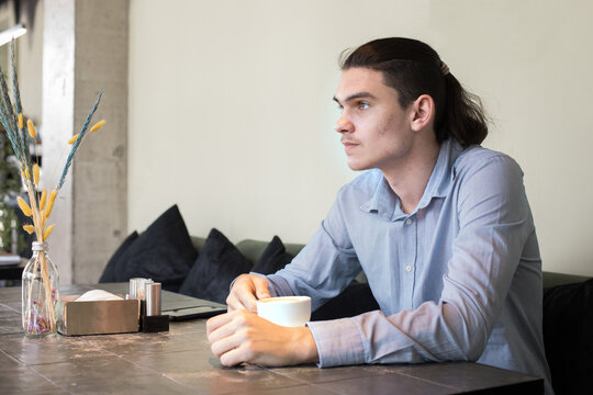 Man sits at table in modern cafe while holding cup and looking out