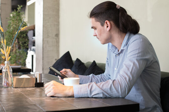 A young man is sitting at a table in a cafe. He looks at his phone and holds a cup in his other hand.