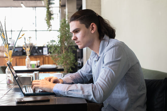 A young man sits at a cafe table focused on his laptop while sipping coffee.