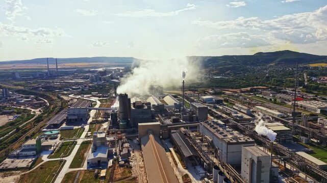 A large factory complex shows multiple buildings and tall chimneys. Smoke rises from the chimneys, filling the air. This scene is set in an industrial area on a clear day.