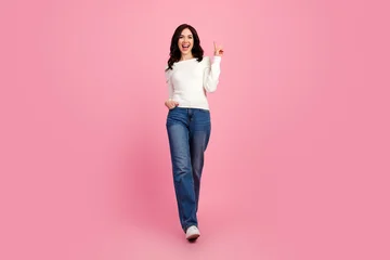 Happy young woman walking and flashing a peace sign in casual jeans and white sweater on a pink background, confident lifestyle portrait and fashion studio shot © deagreez