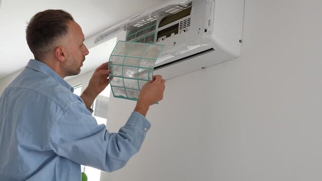 Man removing dirty filter from air conditioner on wall, checking mesh coarse clogged with dust, home appliance maintenance and cooling system care, male inspecting unclean dirty filter from AC blower.
