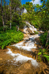 A beautiful natural stream cascades down a limestone cliff and golden rocky slopes at Thararak Waterfall in Mae Sot, Tak, Thailand. Fresh water flows through lush green ferns under a blue sky. © Around Ball