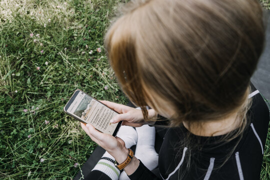 Woman in athletic wear sits on mat in green grass holding smartphone with somatic yoga app instructions. Somatic yoga practice, digital wellness guidance, mindfulness technology, outdoor physical