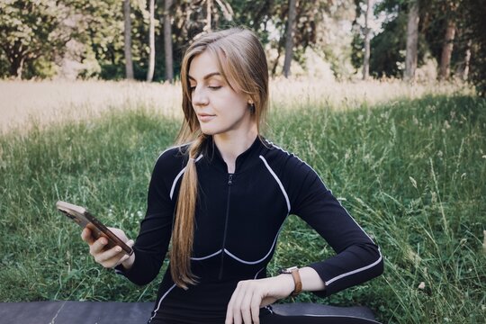 Woman sits on yoga mat in grass looking at smartwatch and holding smartphone. FemTech app tracking, menstrual cycle app, women health technology, women wellness tracker.