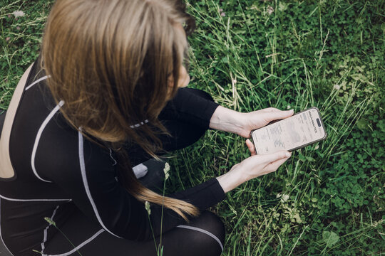 Person sits cross-legged on fitness mat on grass holding smartphone displaying health analytics application. Weekly health summary, wellness data trends, health analytics dashboard, Health report app,