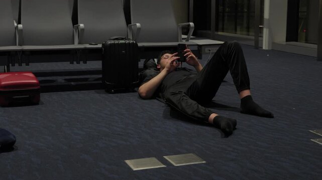 Tired traveler lying on the floor beside luggage using a phone during an overnight layover at an airport terminal