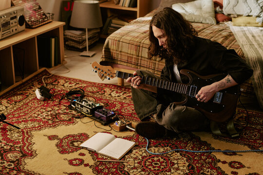 Young adult man playing electric guitar at home, practicing songwriting with effects pedals and notebook, representing music creation. Ideal for creative lifestyle or home studio marketing