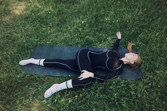 Woman in athletic wear performs supine spinal twist on mat in green grass. Supine spinal twist, reclined twist pose, yoga spine stretch, Lower back release stretch.