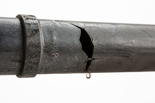 A corroded, cracked metal pipe is dripping a drop of liquid against a white background.