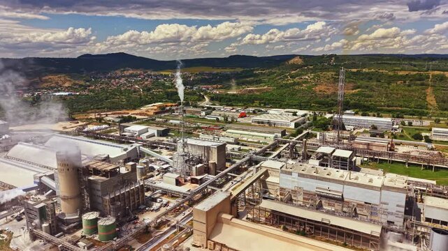 An industrial area with large factories and pipes. Smoke rises from structures under a cloudy sky. Green hills are in the background with some buildings nearby.