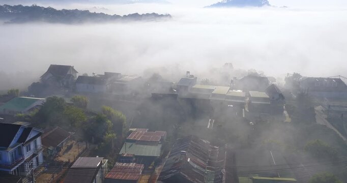 Aerial view of a small hillside village near Dalat, Vietnam wrapped in soft morning fog. Colorful houses emerge through layers of mist, while gentle sunlight illuminates rooftops and greenery.