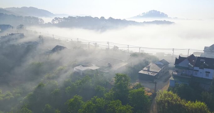 Aerial view of a small hillside village near Dalat, Vietnam wrapped in soft morning fog. Colorful houses emerge through layers of mist, while gentle sunlight illuminates rooftops and greenery.
