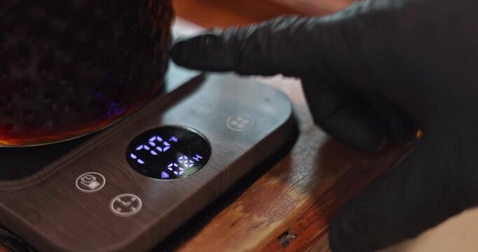 Barista adjusts digital temperature adjustment heater beneath a glass jar, coffee brewing on wooden table
