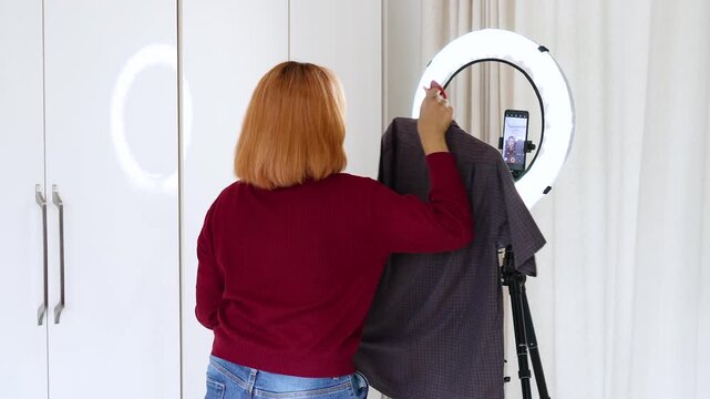 Asian female fashion influencer showing a patterned short-sleeve button-up shirt on a hanger while live streaming or recording a video with a ring light and smartphone at home.