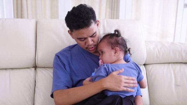 Caring Asian father gently patting the back of his sleepy toddler daughter while holding her closely on a living room sofa.