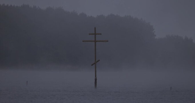 Close up view of an orthodox wooden cross standing in a lake in thick fog