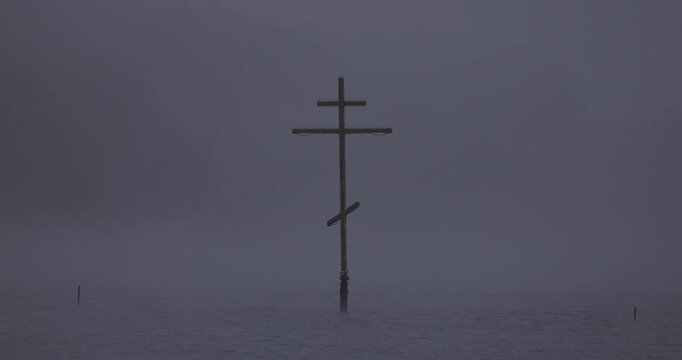 Close up view of an orthodox wooden cross standing in a lake in thick fog