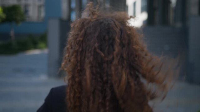 Happy Caucasian business woman turning back touching curly hair enjoying shampoo result walking in city near office building. View from behind businesswoman female girl enjoy hairstyle walk outdoors