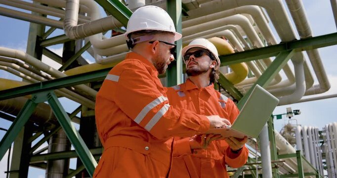 Adult male technician types on laptop while male supervisor reviews refinery pipeline layout, coordinating petrochemical maintenance planning, safety compliance audit, operation monitoring review