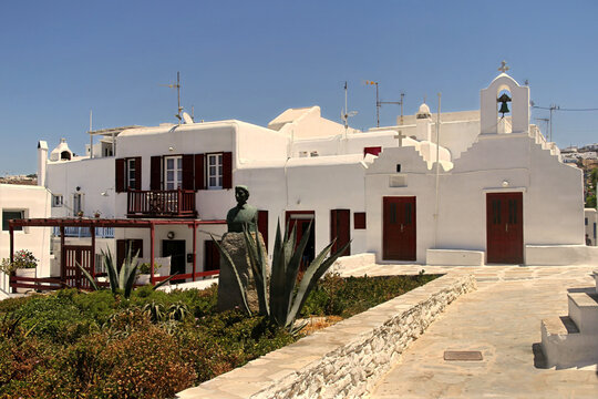 A scenic square in Mykonos Town featuring the historic bust of local war heroine Manto Mavrogenous against traditional whitewashed buildings.