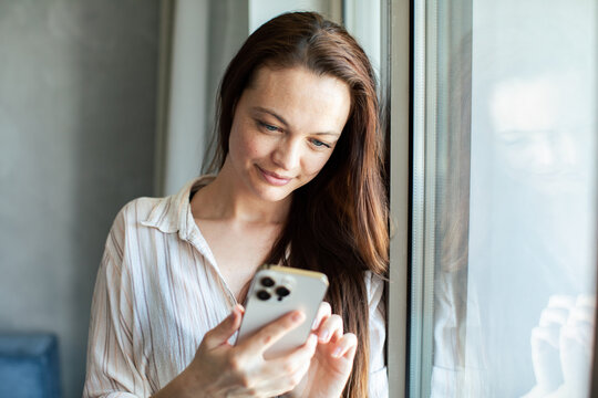 Smiling woman texting on smartphone by window at home