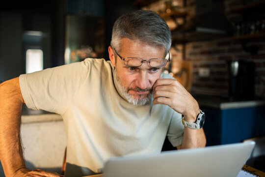 Mature man working on laptop at home kitchen