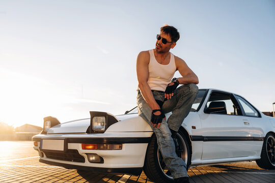 Male car enthusiast in sleeveless shirt and gloves poses on white sports car during sunset, showcasing vehicle's sleek design and urban backdrop