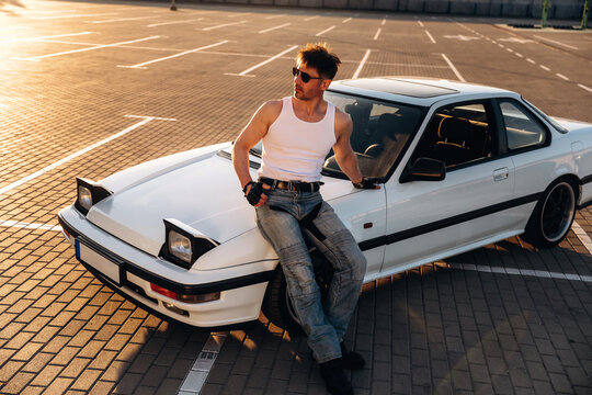 Male car enthusiast in sleeveless shirt and sunglasses sits on white sports car in empty parking lot during sunset, showcasing vehicle's sleek design and surroundings