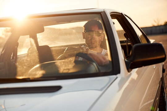 Male driver wearing sunglasses sits inside a white car with sunlight streaming through the windshield, showcasing a scenic outdoor environment in the background