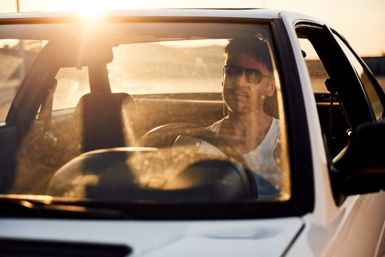 Male driver wearing sunglasses sits in a white car with sunlight streaming through the windshield, creating reflections and a warm glow inside the vehicle
