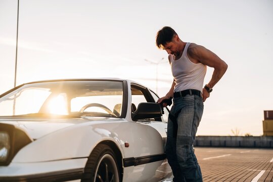 Male mechanic in sleeveless shirt and jeans checks car door handle of white sports car parked on rooftop during sunset with city skyline in the background