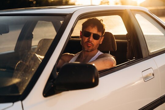 Male driver wearing sunglasses leans against the window of a white car during sunset, showcasing a relaxed demeanor in a warm outdoor setting