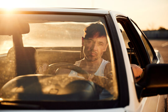 Male driver in sleeveless shirt sitting in white car with sunlight reflecting off the windshield, parked in an outdoor setting during golden hour