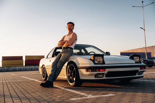 Caucasian male car enthusiast in sleeveless shirt poses confidently beside a vintage sports car parked on a sunlit outdoor pavement with shipping containers in the background