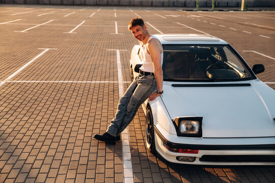 Caucasian male leaning against a white sports car in a parking lot, wearing a sleeveless shirt and jeans, with a sunny sky and empty parking spaces in the background