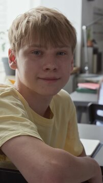 Portrait of Caucasian teenage boy sitting at desk in classroom, looking at camera and smiling. Footage for educational marketing, school promotions, and e-learning campaigns, vertical