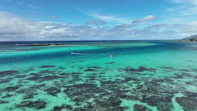 Kitesurfer glides across turquoise lagoon near Tiahura, Moorea, French Polynesia, revealing dark coral reef patterns beneath clear water, distant green mountain coast. Aerial drone view.